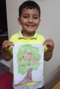 Chlapec ukazuje pastelkami kreslený obrázek stromu.
