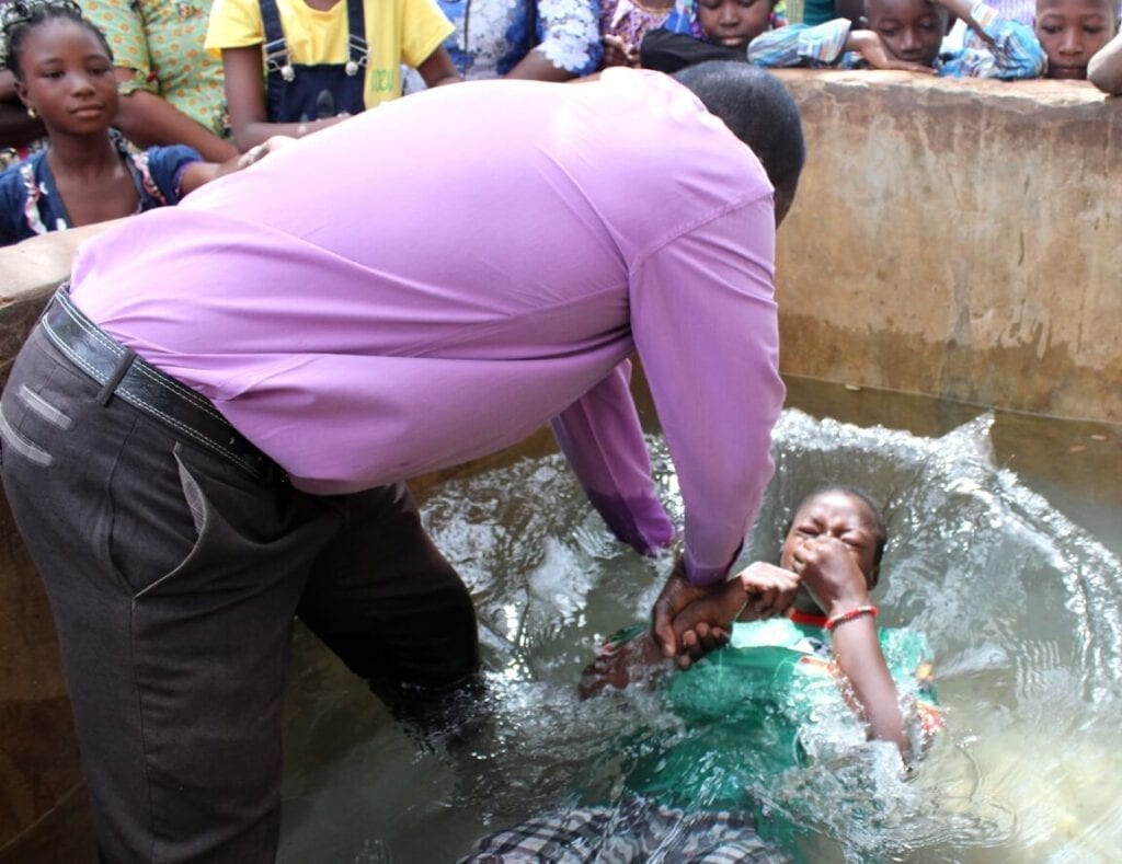 Muž křtí školáka v nádrži s vodou, spolužáci přihlížejí.