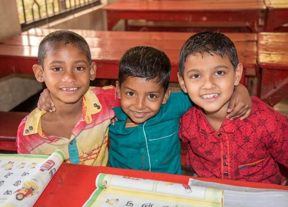 Tři malí spolužáci ve třídě se v přátelském gestu drží za ramena. Díky adopci dětí na dálku se mohou učit.